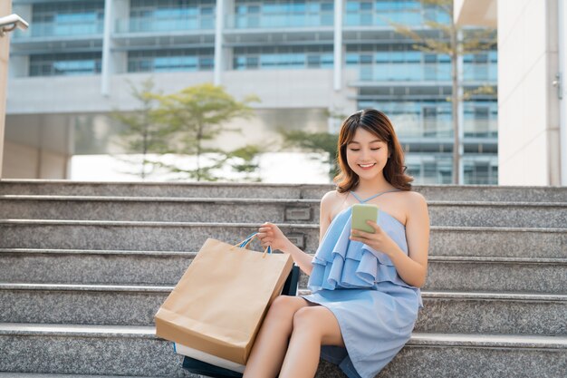 電話を使用して、屋外の階段に座っている買い物袋を持つ美しい若い女性