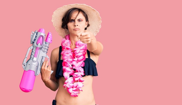 Beautiful young woman with short hair wearing bikini and hawaiian lei holding water gun pointing with finger to the camera and to you, confident gesture looking serious