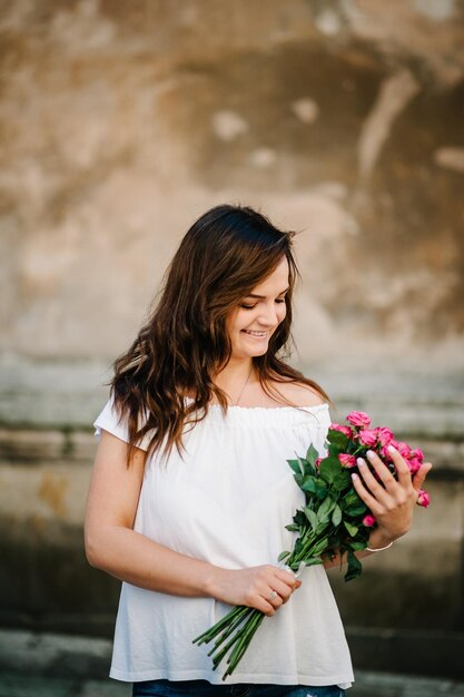 Beautiful young woman with spring flowers bouquet at city street Happy girl smiling and holding pink roses flowers outdoors Spring portrait of pretty female