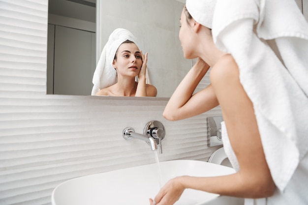 Bella giovane donna con un asciugamano sulla testa in piedi in bagno, esaminando il suo viso in uno specchio