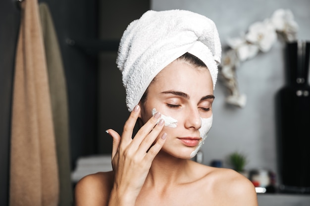 Bella giovane donna con un asciugamano avvolto intorno alla sua testa che applica la maschera per il viso in bagno