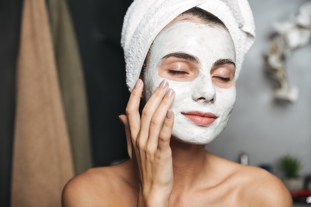 Beautiful young woman with towel wrapped around her head applying face mask at the bathroom