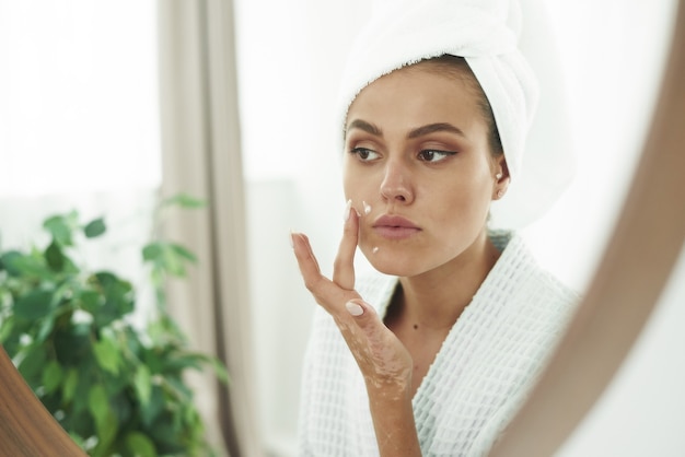 Una bella giovane donna con la vitiligine sulle mani in accappatoio si applica una crema sul viso