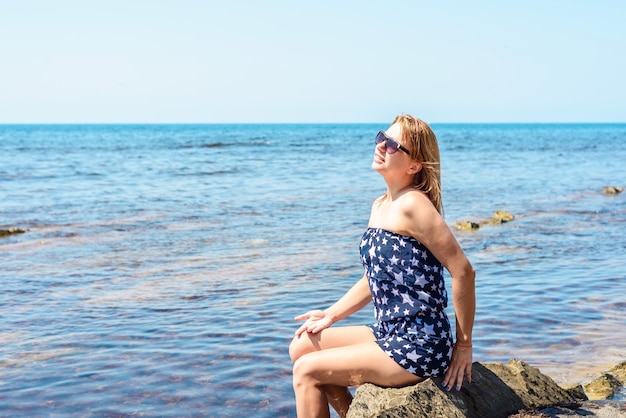 Beautiful young woman with a white hut is sitting on a stone on Lalaria Beach Skiathos island.