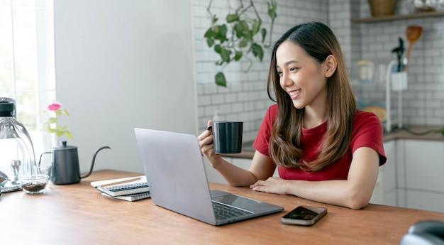 Beautiful young woman working on laptop computer while sitting at the kitchen room drinking coffee