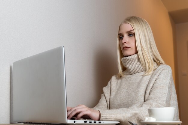 リビングルームに座って、コーヒーを飲みながらラップトップコンピューターで作業している美しい若い女性。