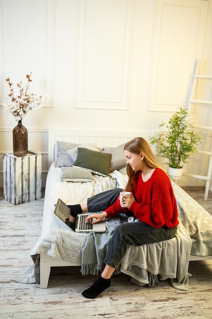 Beautiful young woman working on a laptop sitting on the bed at home, she is drinking coffee and smiling. Work from home during quarantine. Girl in a sweater and jeans at home on the bed