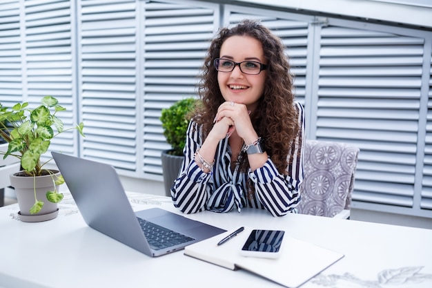 Bella giovane donna che lavora su un computer portatile e sorride mentre è seduta all'aperto in un caffè. giovane donna che utilizza computer portatile per lavoro. libero professionista femminile che lavora su un computer portatile in un caffè all'aperto.