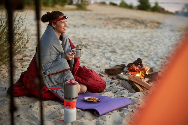 Beautiful young woman wrapped in sleeping bag holding cup of tea and smiling while sitting on sand near campfire