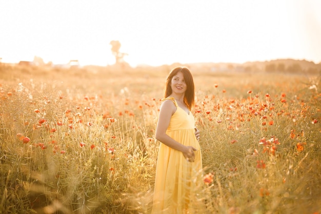 Beautiful young woman in a yellow dress walking in a poppy field on a summer day. Girl enjoying flowers in the countryside. Selective focus