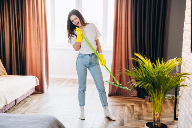 Красивая молодая женщина в желтых перчатках поет со шваброй во время уборки в гостиной.