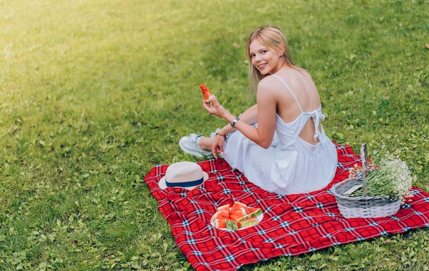 格子縞の上に座って、スイカを食べる美しい若い女性。自然、ピクニック。