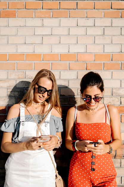 Beautiful young women texting on mobile phones while standing near brick wall on sunny day