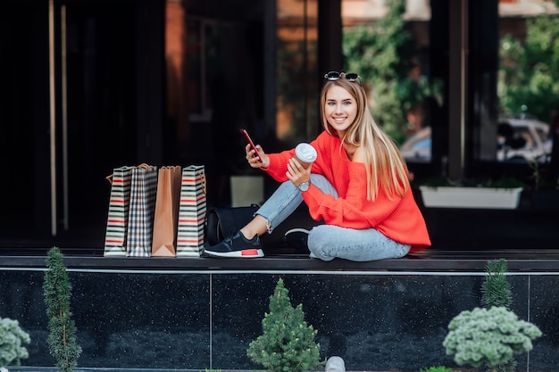 Beautifulblonde woman sitting on street and surrounded by shopping bags and looking on phone. Fashion street concept.