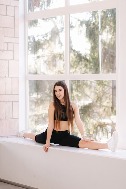 Beautifull brunette woman doing twine stretching on windowsill at home. Attractive fit girl training at home. Copy space.
