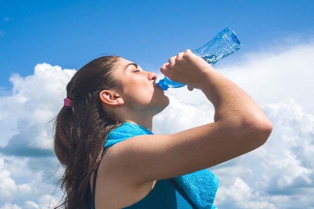 Красивая бегунья девушка имеет перерыв, пьет воду против ясного голубого неба.