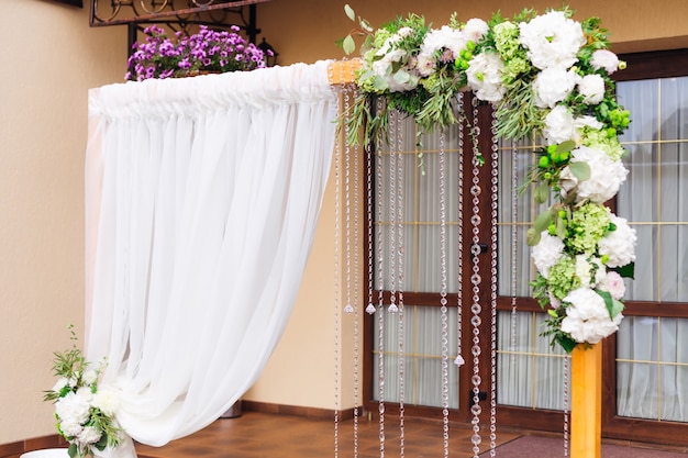 Cerimonia di visita splendidamente decorata vicino alle porte in vetro del ristorante arco in legno con composizione floreale in tulle e catene con perline trasparenti