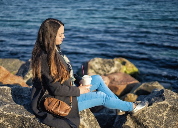 テイクアウトコーヒーと岩のビーチに座っているBeautigulの女の子