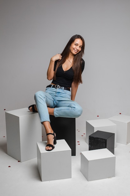 Beautiufl caucasian woman with charming appearance sits on a white cube and looks, picture isolated on white wall