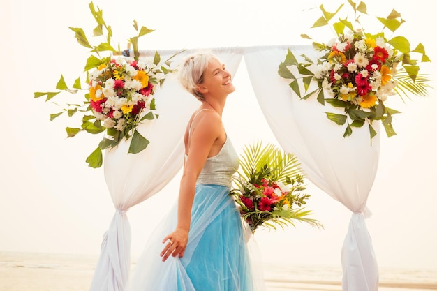 Beautuful blonde kortharige vrouw poseren in blauwe tule lange trein jurk in de buurt van bruiloft bloem boog met tropisch boeket op zandstrand