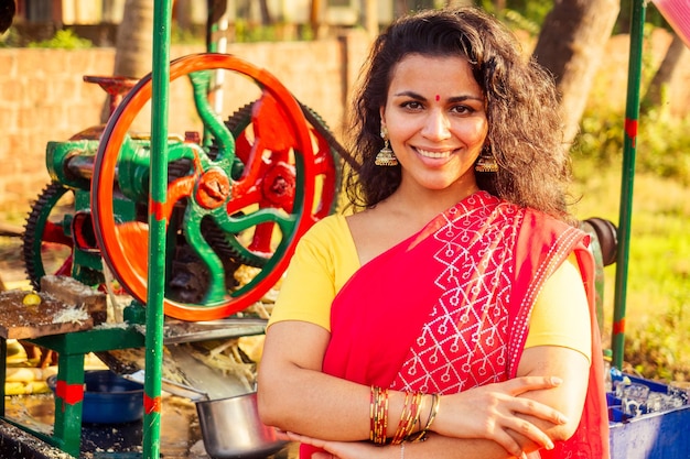 Beautuful induan woman in red sari standing near sugarcane juice maker apparatus machine plantation summer farm background small business start up