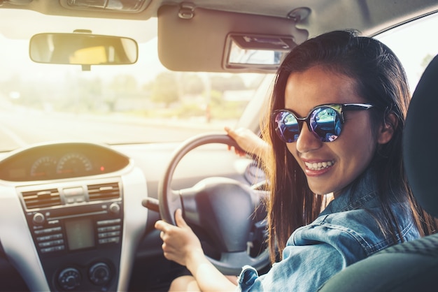 Sorriso asiatico della donna di bellezza con la giacca del tralicco blu dei sunglass che conduce automobile al tempo di tramonto.