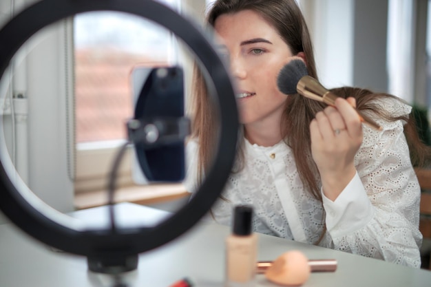 Beauty blogging technology and people concept portrait of a happy smiling girl blogger with ring light and smartphone applying make up at home making a influencer video