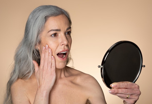 Beauty care shocked senior woman looking in magnifying mirror with open mouth enjoying new treatment