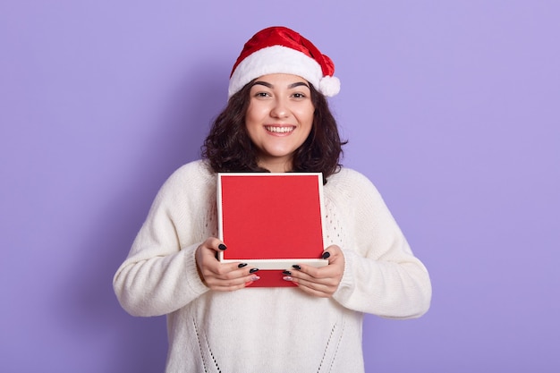 Beauty christmas fashion model meisje met rode geschenkdoos, direct kijken, heeft donker golvend haar en mooie manicure, meisje met witte gebreide trui en kerstmuts.