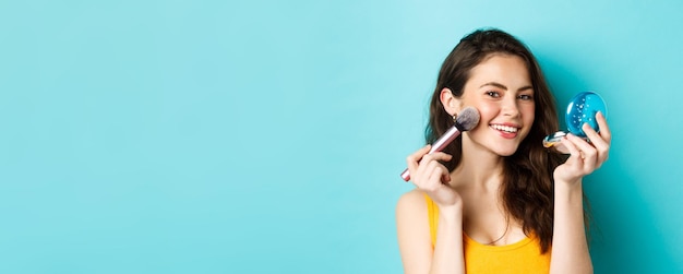 Beauty Close up of attractive woman looking in mirror and using brush to apply make up smiling pleased at camera standing against blue background