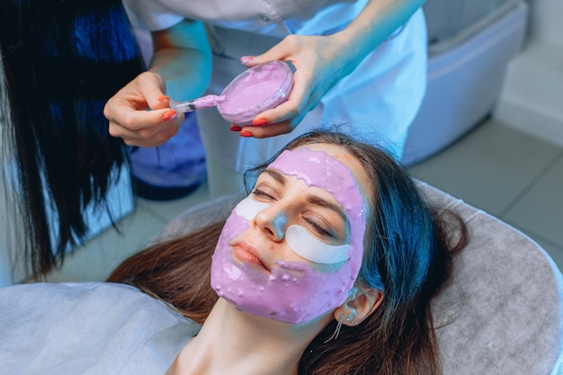 Beauty complex of facial skin care procedures. A cosmetologist makes an alginate mask for a girl in fabric patches under her eyes.