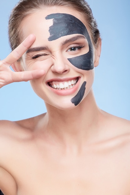 Beauty concept, head and shoulders of girl with grey mask on face, face pack. Closeup of young woman smiling broadly and showing V, One eye closed. Studio, grey background