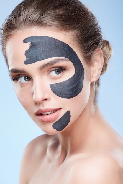 Beauty concept, head and shoulders of girl with grey mask on half face, face pack. Young woman with pure skin looking aside. Studio, grey background