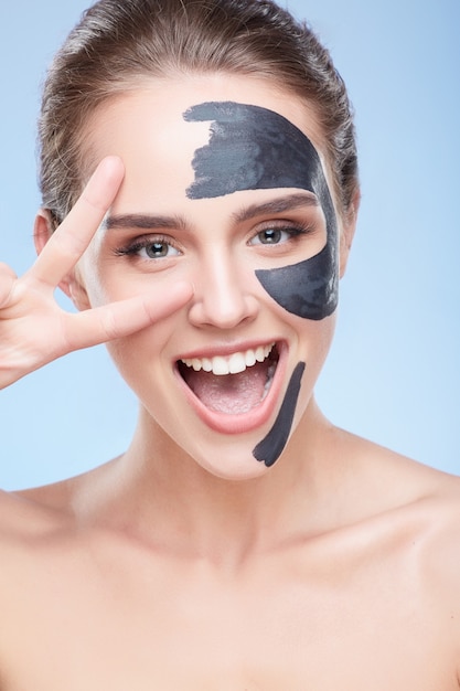 Beauty concept, head and shoulders of smiling girl with grey mask on face, face pack. Closeup of delighted woman showing V, smiling and looking at camera. Studio, grey background