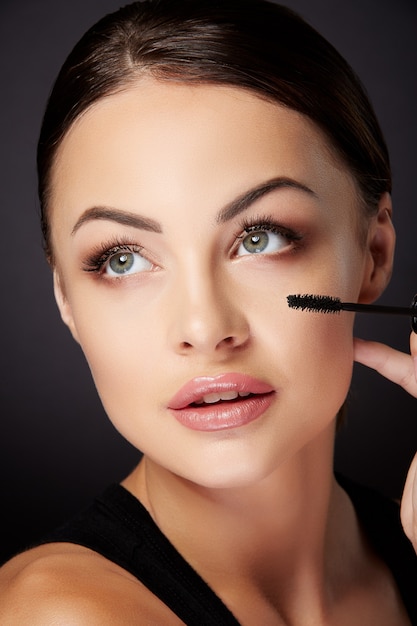 Beauty concept, woman holding mascara. Beauty portrait of woman looking up, painting lashes with mascara. Studio, inside, black background