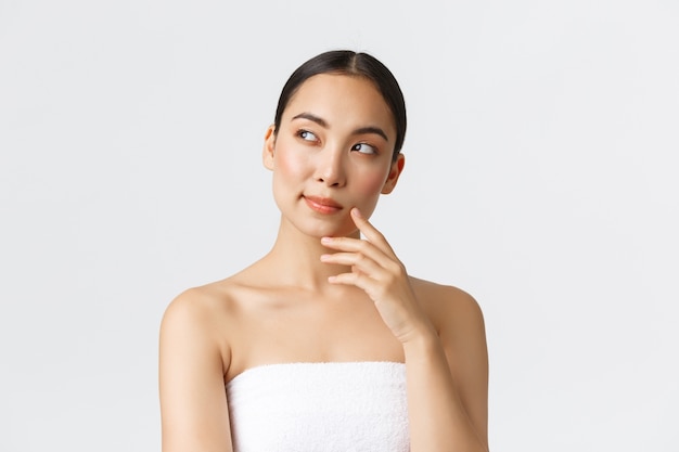 Beauty, cosmetology and spa salon concept. Thoughtful beautiful asian woman in towel looking left and thinking, pondering or making choice which procedure take in beauty clinic.