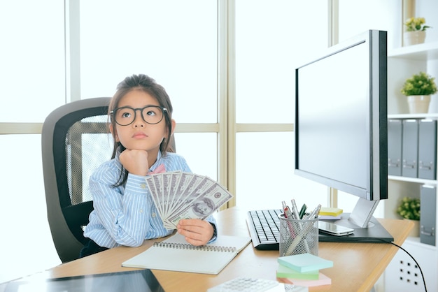 beauty cute little girl kid play as business people sitting on working desk and holding banknote thinking how to saving money.