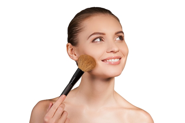 Beauty doing make-up. Beautiful young shirtless woman holding make-up brush near face and smiling while standing isolated on white background