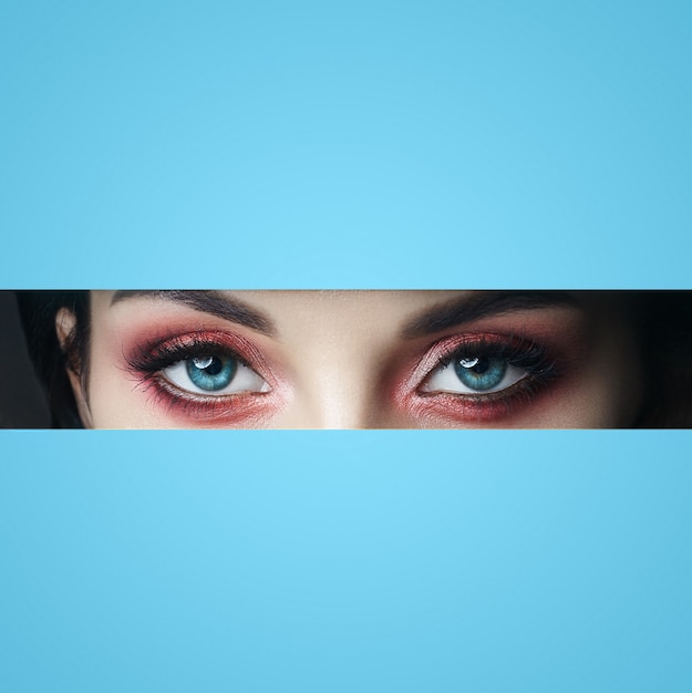 Beauty face red makeup eyes of a young girl in a slit hole of\
blue paper. woman with beautiful makeup red glowing shadow, big\
blue eyes in the slit hole