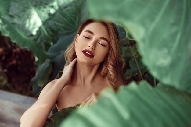 Photo beauty face. woman model with bright makeup and healthy skin behind green leaf plant
