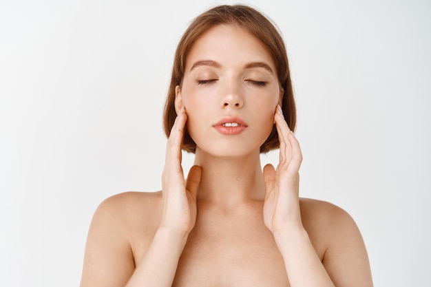 beauty face. young woman rubbing creme in cheeks. girl with healthy hydrated facial skin, close eyes and gently youching natural face without make up, standing on white wall