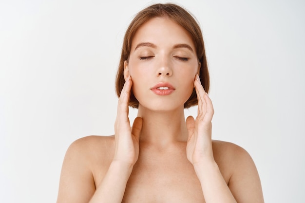 Beauty face. Young woman rubbing creme in cheeks. Girl with healthy hydrated facial skin, close eyes and gently youching natural face without make up, standing on white wall