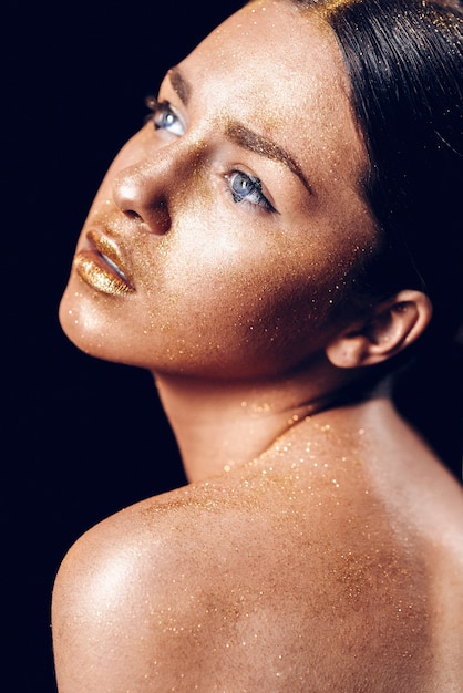 Beauty fashion art model girl with Golden skin. Close up portrait of girl with blue eyes on black background