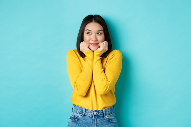 Beauty and fashion concept. Beautiful asian woman blushing and smiling, looking dreamy left, imaging something cute, standing against blue background.