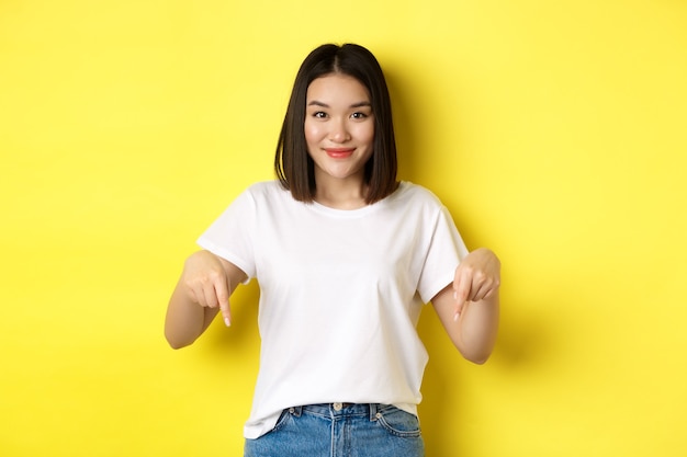 Beauty and fashion concept. Beautiful asian woman in white t-shirt pointing fingers down, demonstrate logo standing over yellow background.