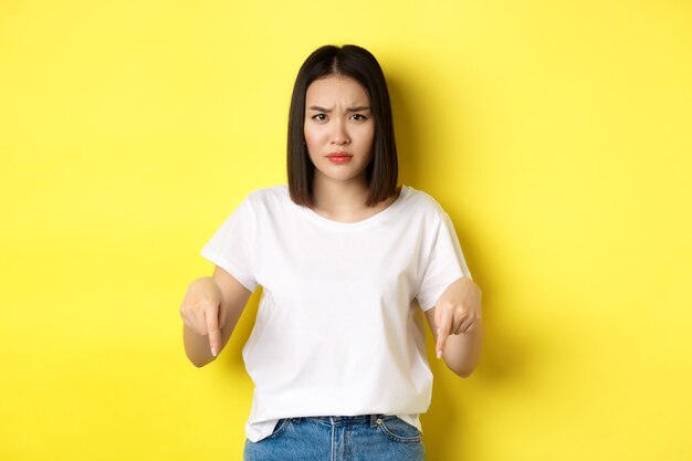 美容とファッションのコンセプト。指を下に向けて白いTシャツを着た美しいアジアの女性は、黄色の背景の上に立っているロゴを示しています。
