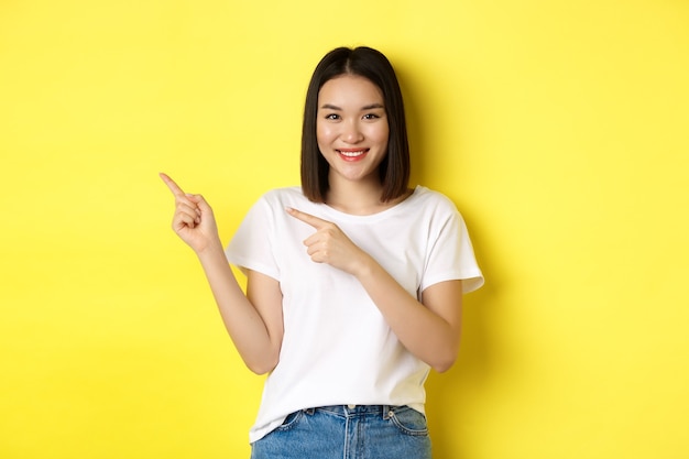 美容とファッションのコンセプト。黄色の背景の上に立って、左の指を指す白いTシャツの美しいアジアの女性。
