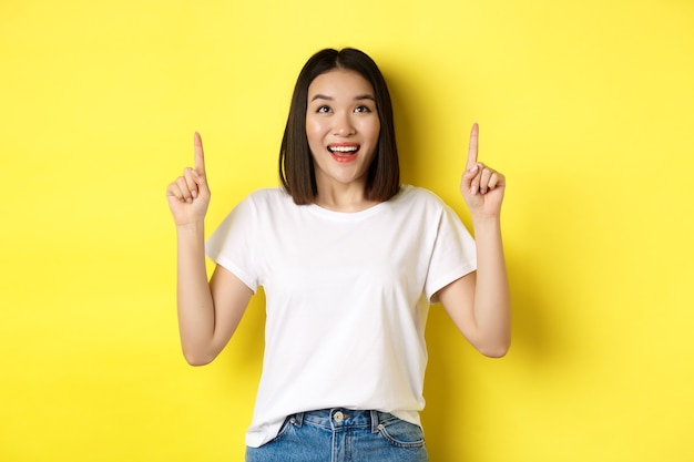 Beauty and fashion concept. Beautiful asian woman in white t-shirt pointing fingers up, standing over yellow background