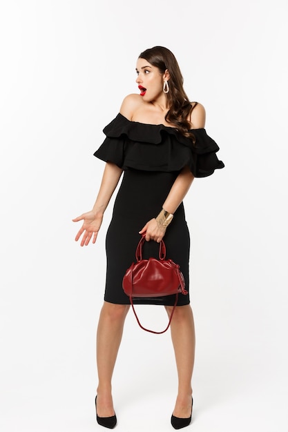 Photo beauty and fashion concept. full length of surprised woman in elegant dress, heels looking left confused, holding purse, cant understand what happening, white background