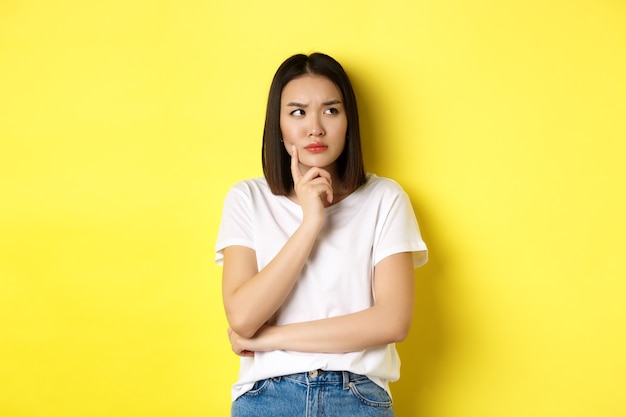 Beauty and fashion concept. Pensive asian woman thinking, looking thoughtful while pondering something, standing over yellow background.